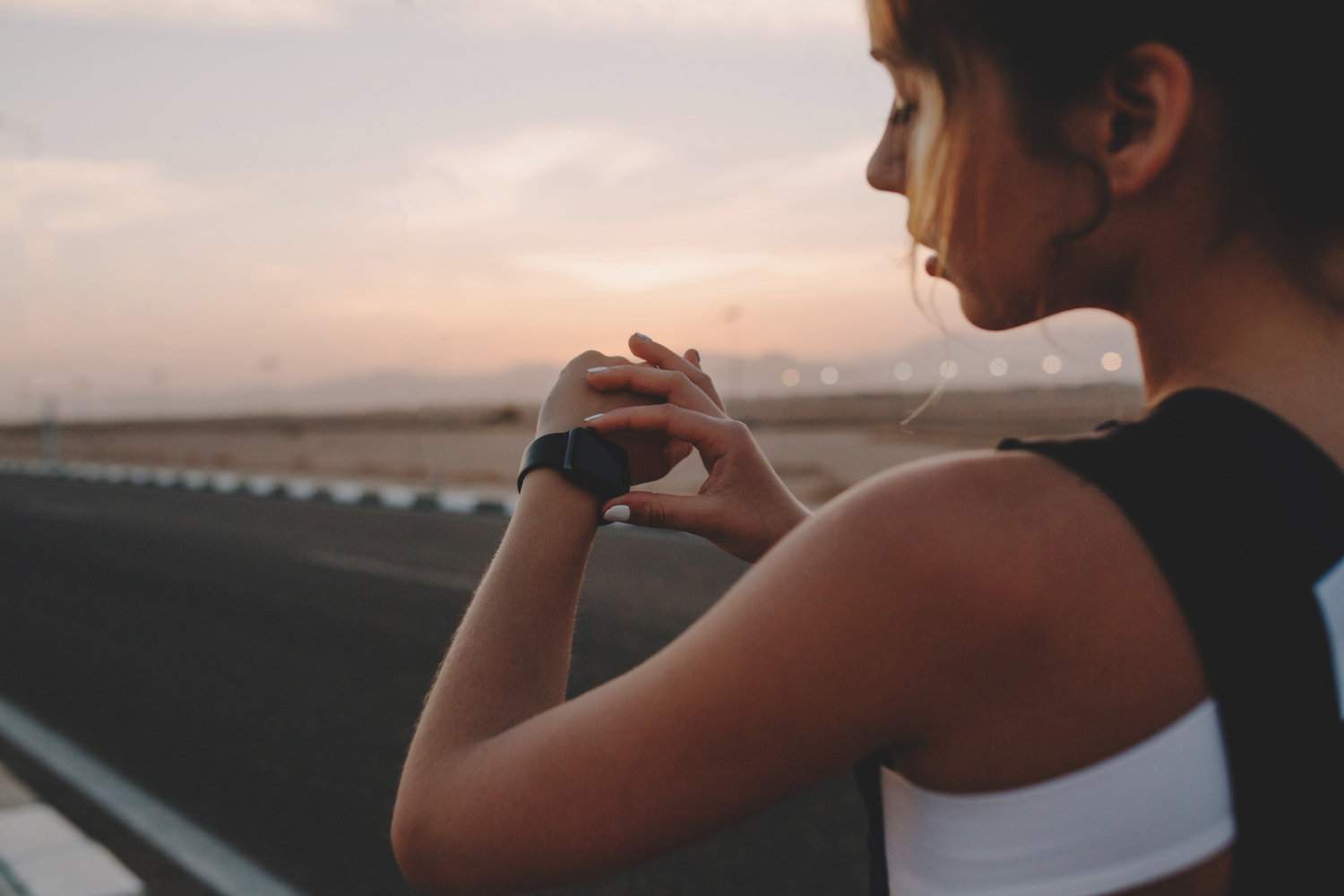 A woman in workout clothes stands beside an empty road at dusk, looking down at a smartwatch on her wrist before exercising.