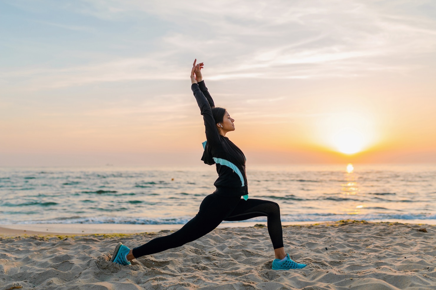 A woman holds a yoga lunge pose on a sandy beach at sunrise, with both arms extended overhead toward the sky.