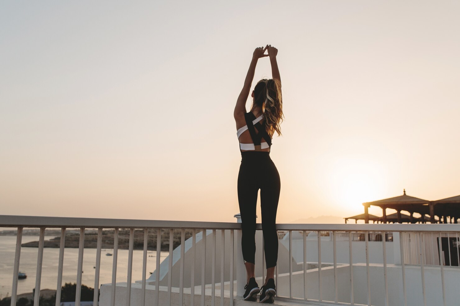 A woman stands on a terrace overlooking the water at sunrise, stretching both arms overhead in black workout clothes.