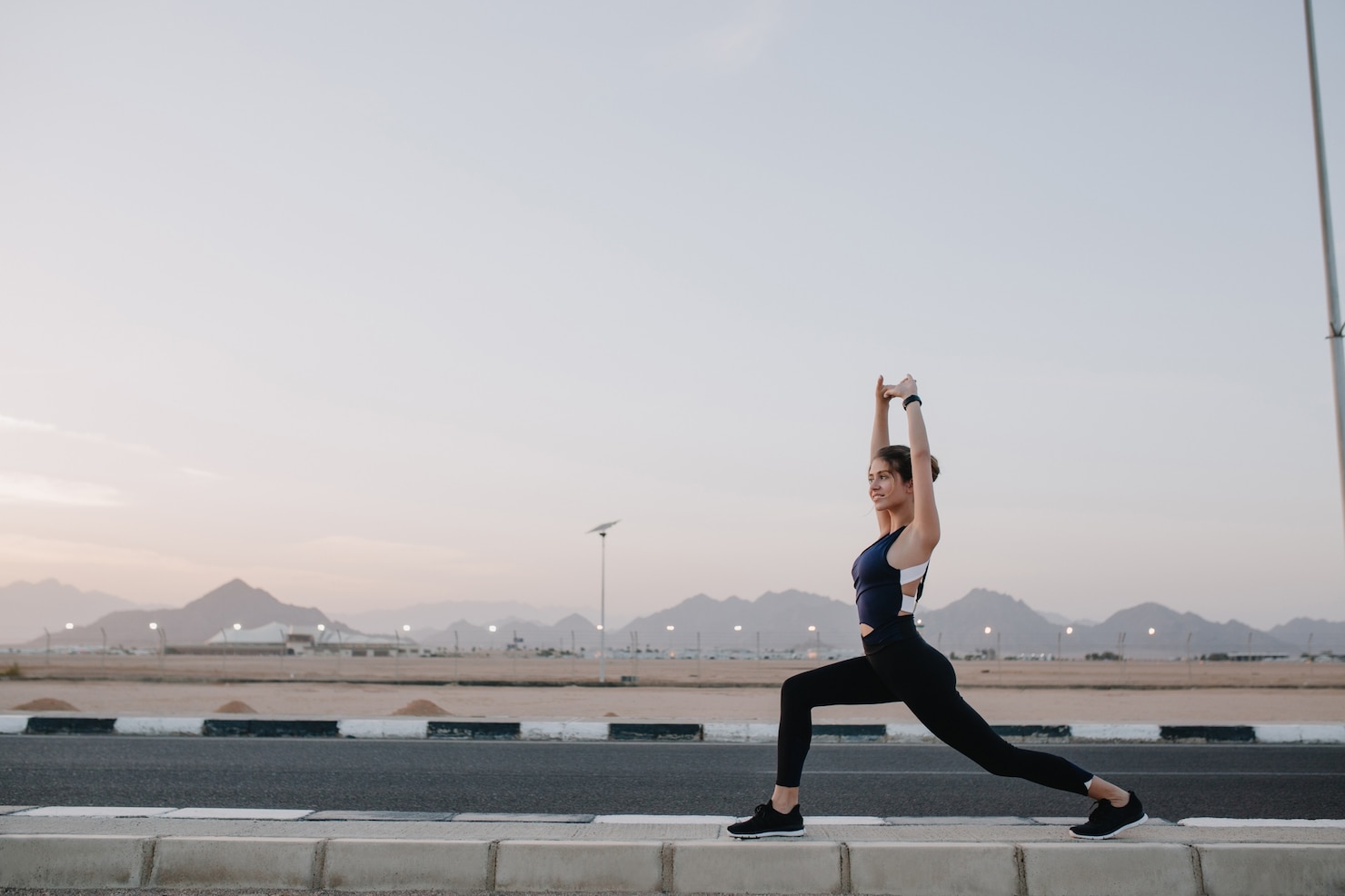 A woman holds a deep lunge stretch on a curbside road at dawn, with mountains and an open sky behind her.