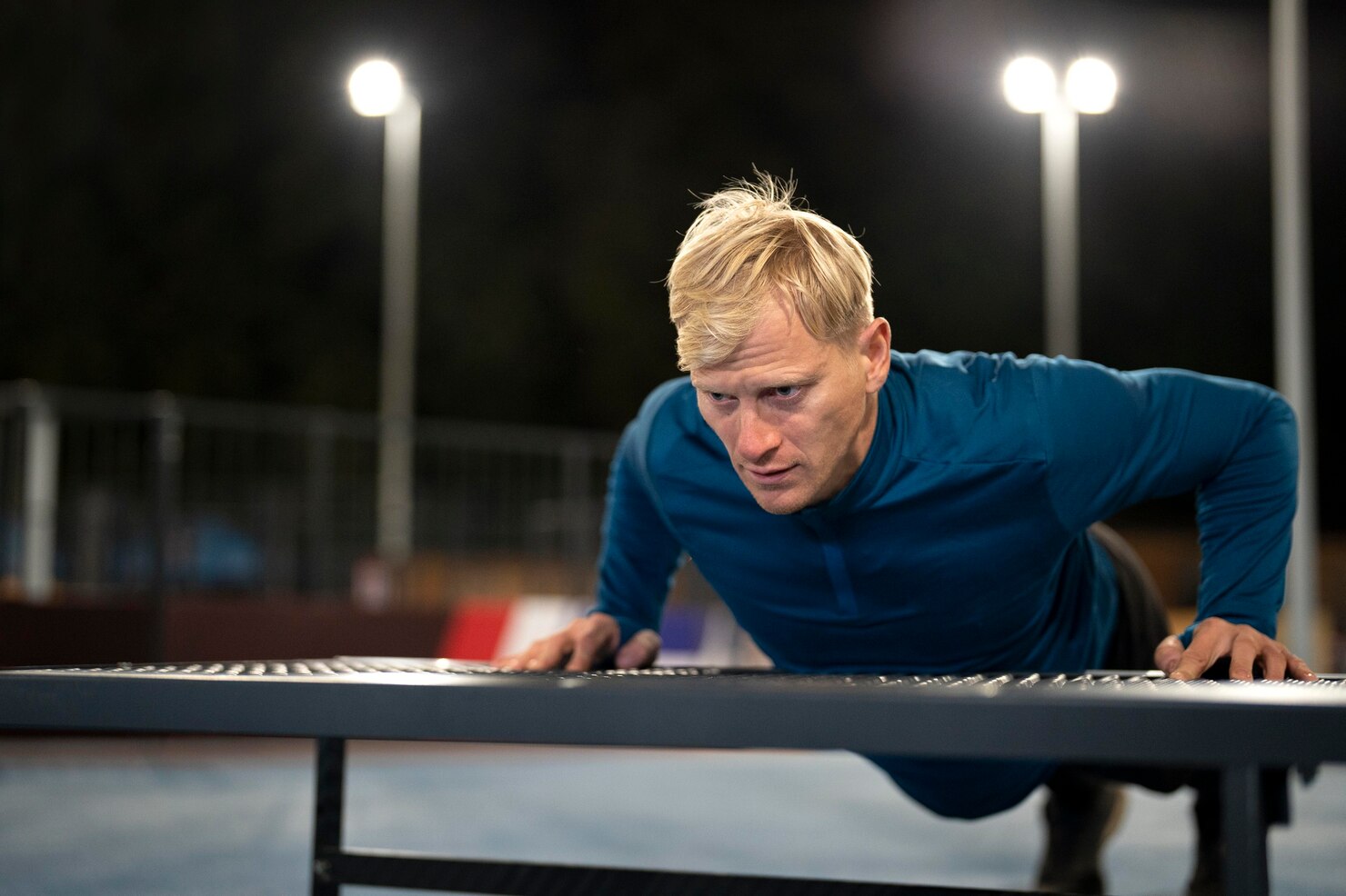 A man in a blue long-sleeve top does pushups on an outdoor bench at night under bright court lights.