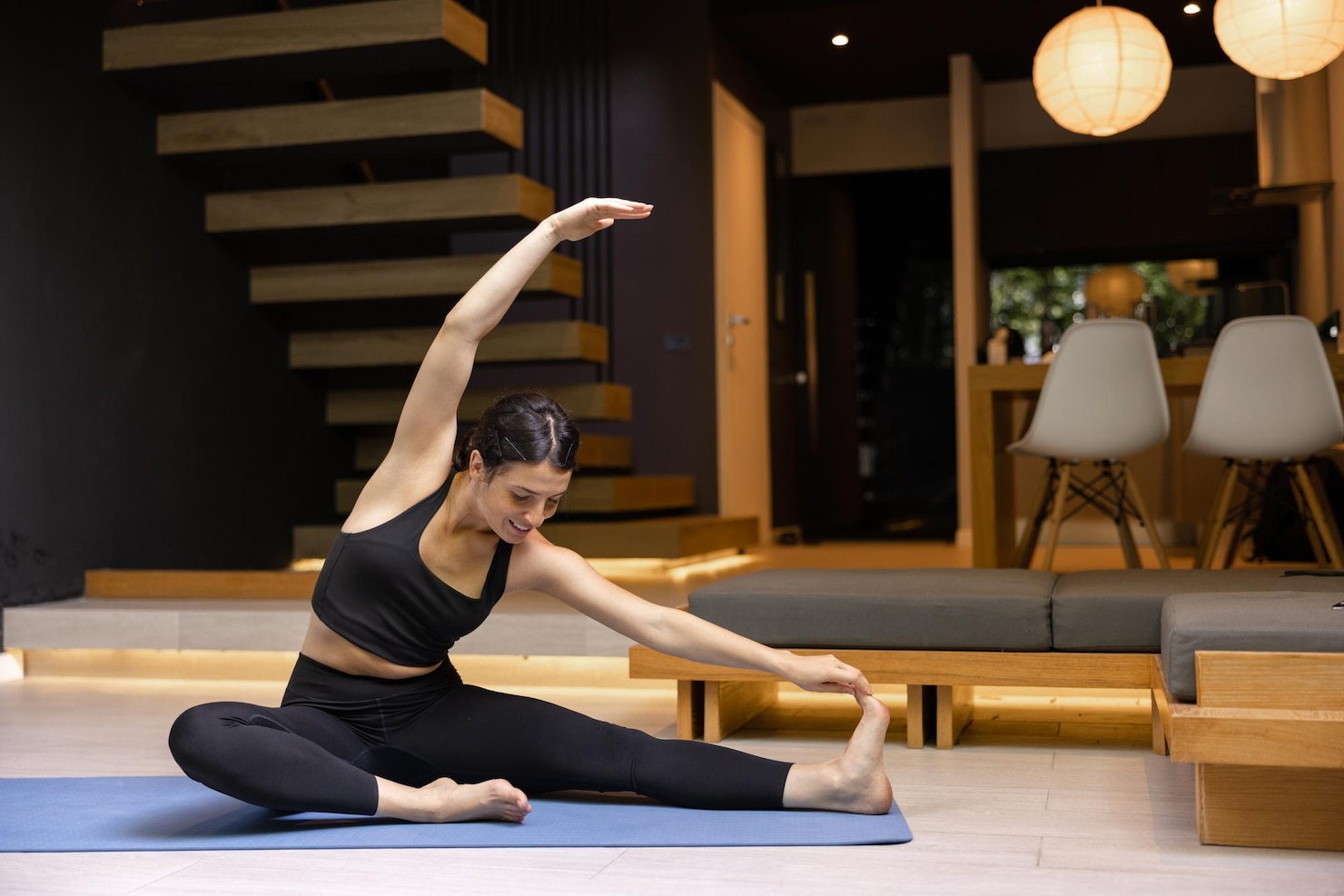 A woman sits on a yoga mat in a modern living room, reaching one hand toward her foot while stretching with the other arm curved overhead.