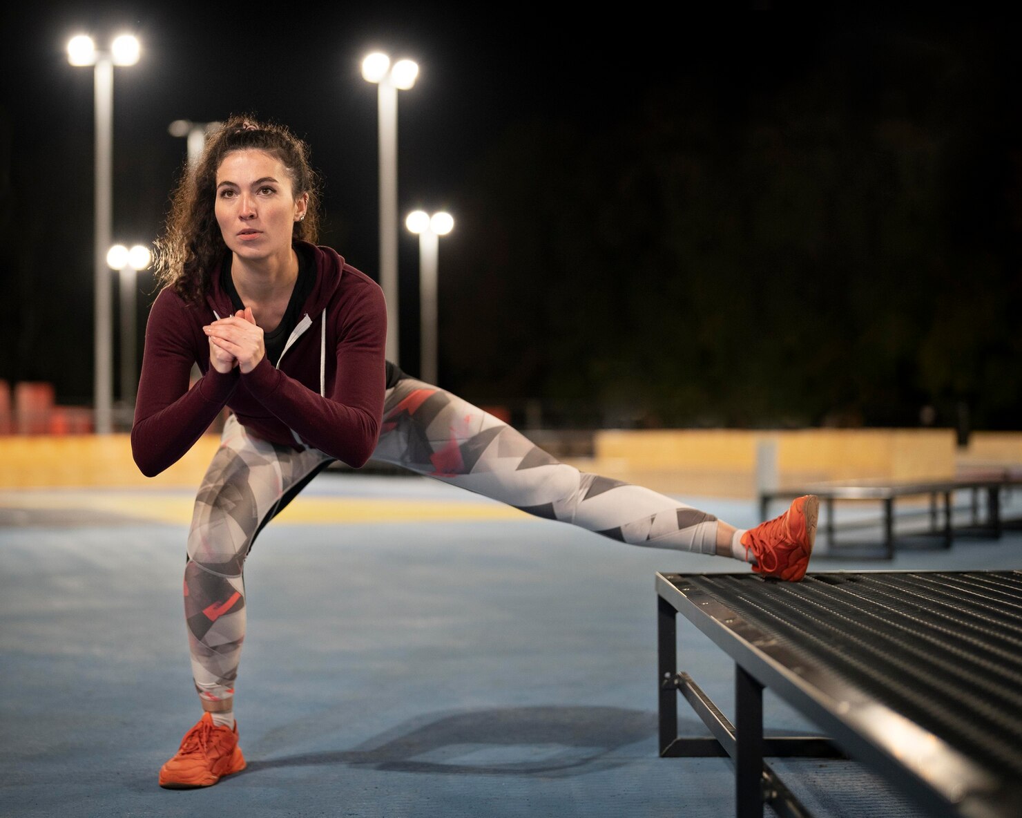 A woman in athletic clothes performs a side lunge stretch with one foot raised on a bench at an outdoor court lit at night.
