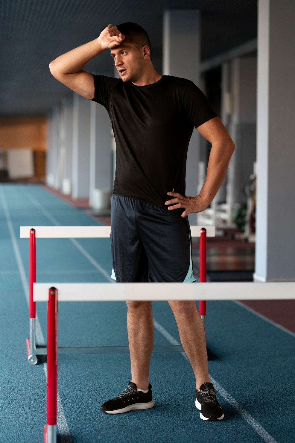 A man in black athletic wear stands on an indoor track beside hurdles, wiping sweat from his forehead after a workout.