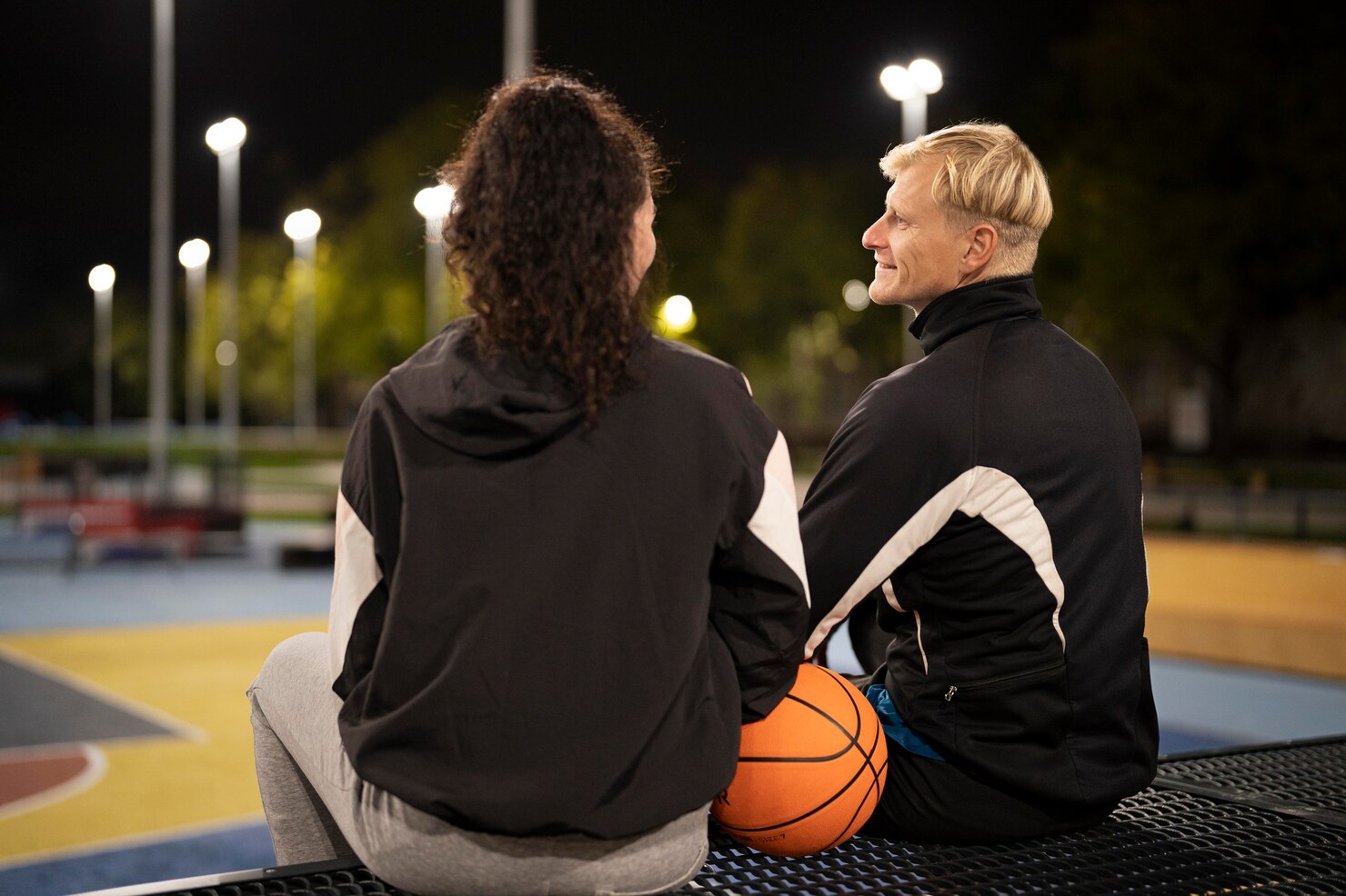 Two people sit together on a bench at an outdoor court at night, talking beside a basketball under tall floodlights.