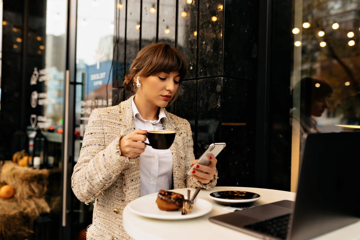 A woman sits at a small cafe table holding a black coffee cup in one hand and checking her phone with the other. A pastry plate and laptop sit on the table in front of her.
