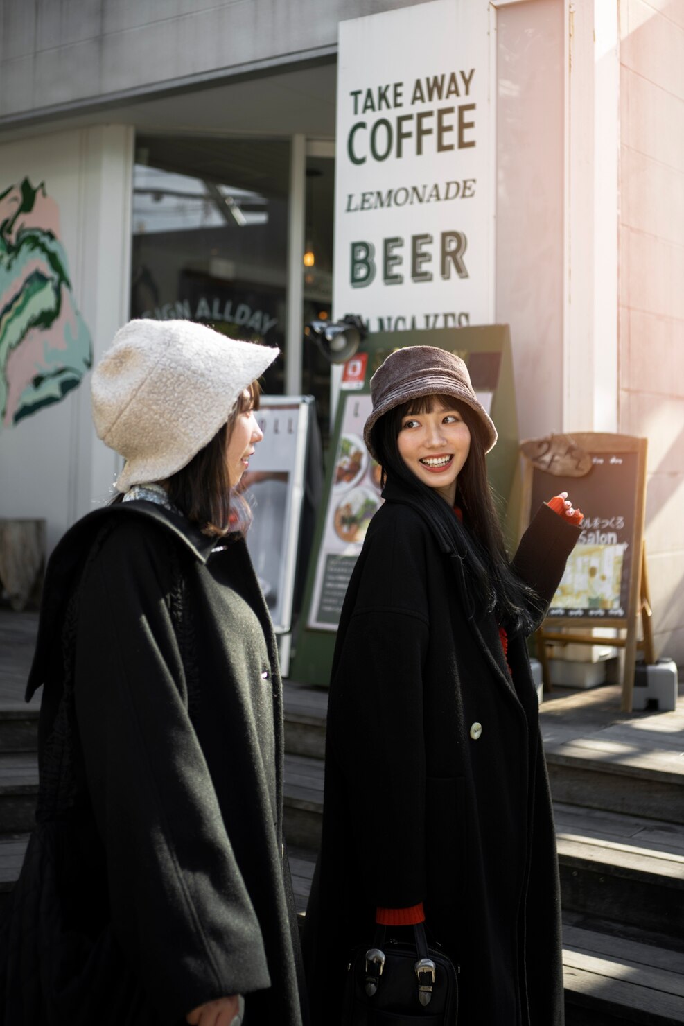 Two women in bucket hats walk past a cafe entrance with a large sign advertising takeaway coffee, lemonade, and beer. One of them looks back smiling toward her companion.