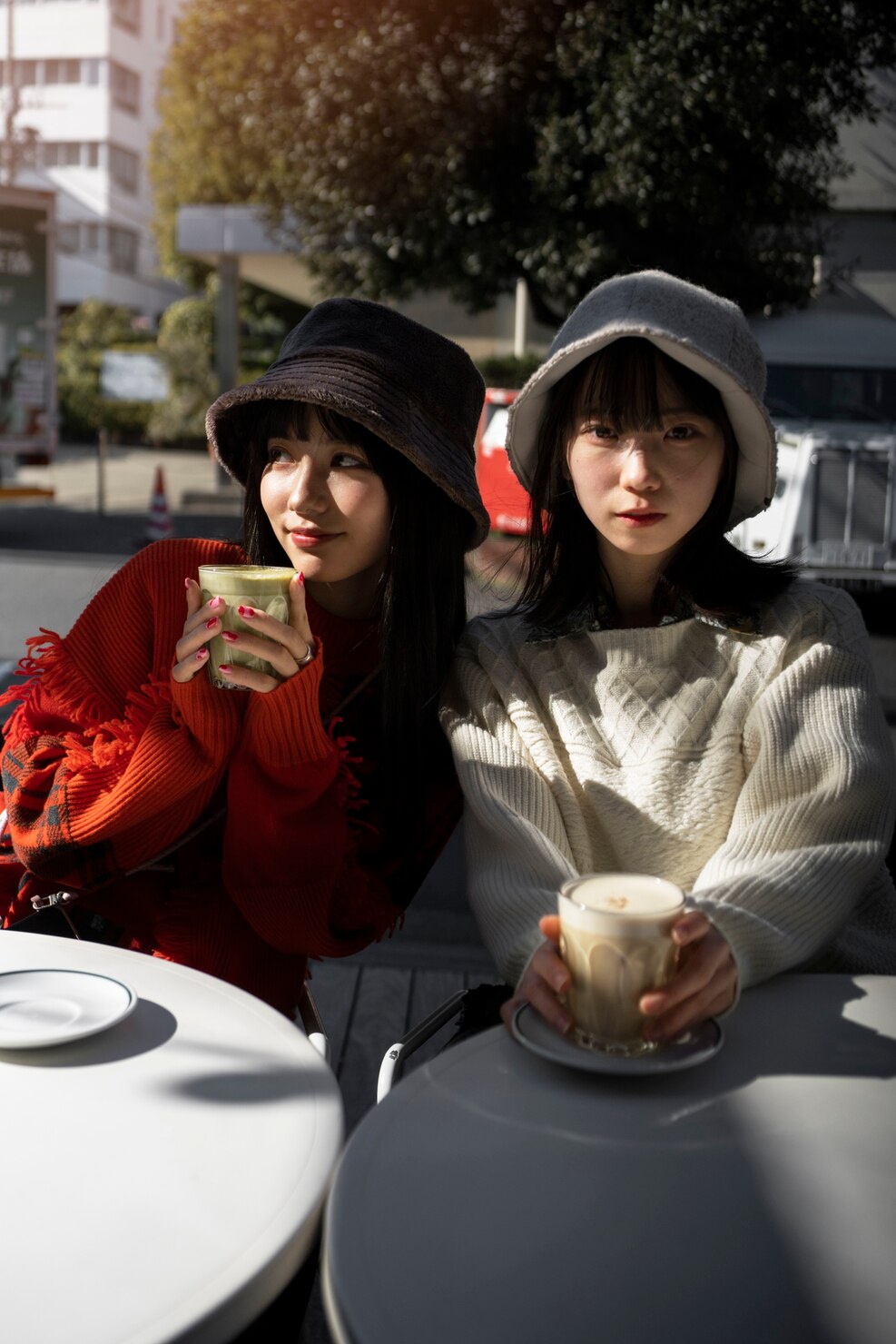 Two women sit outdoors at cafe tables holding creamy coffee drinks in glass cups. One wears a red sweater and dark hat, while the other wears a light sweater and pale bucket hat.