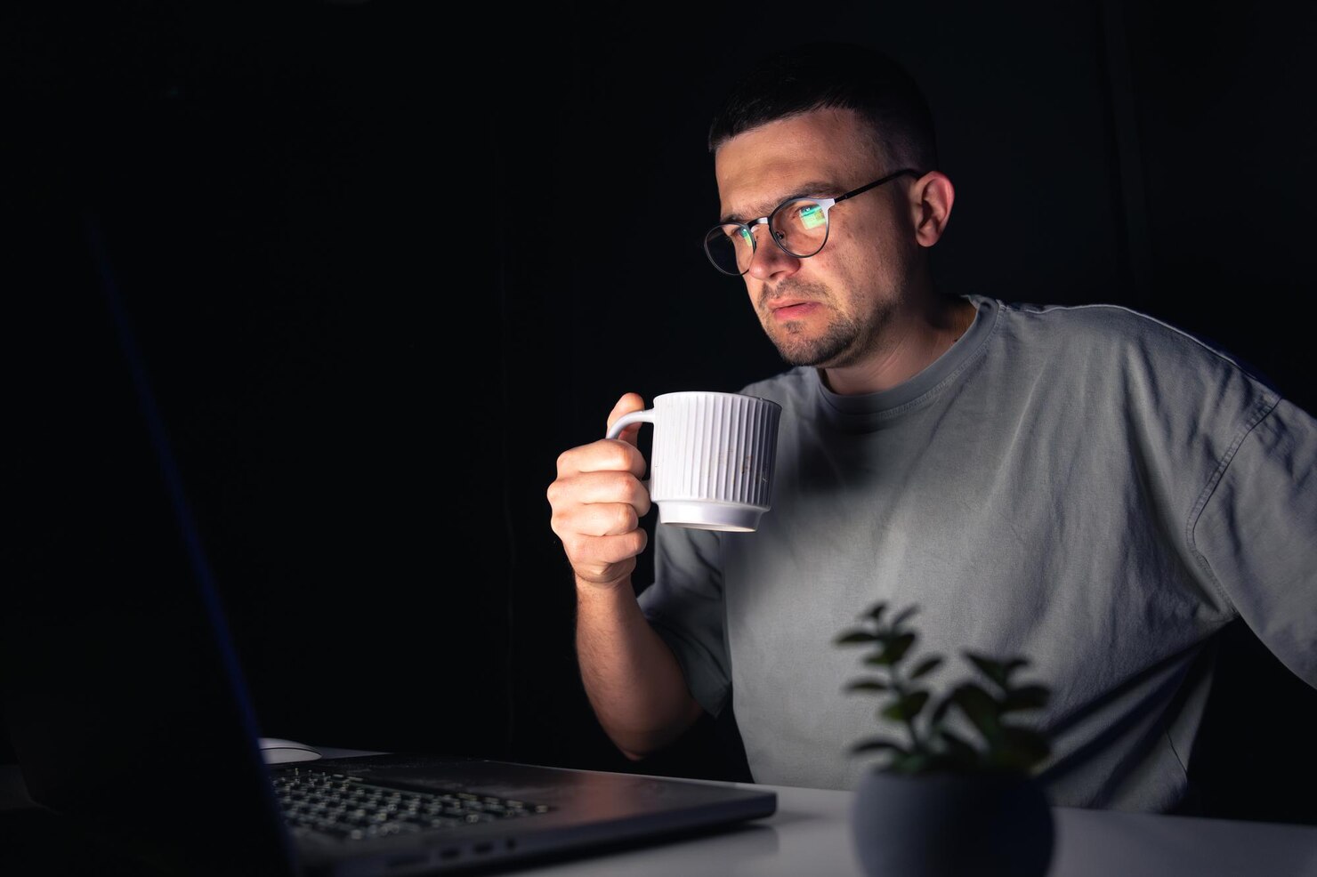 A man in glasses sits in a dark room holding a large white mug while looking toward a laptop screen, lit mainly by the screen glow.