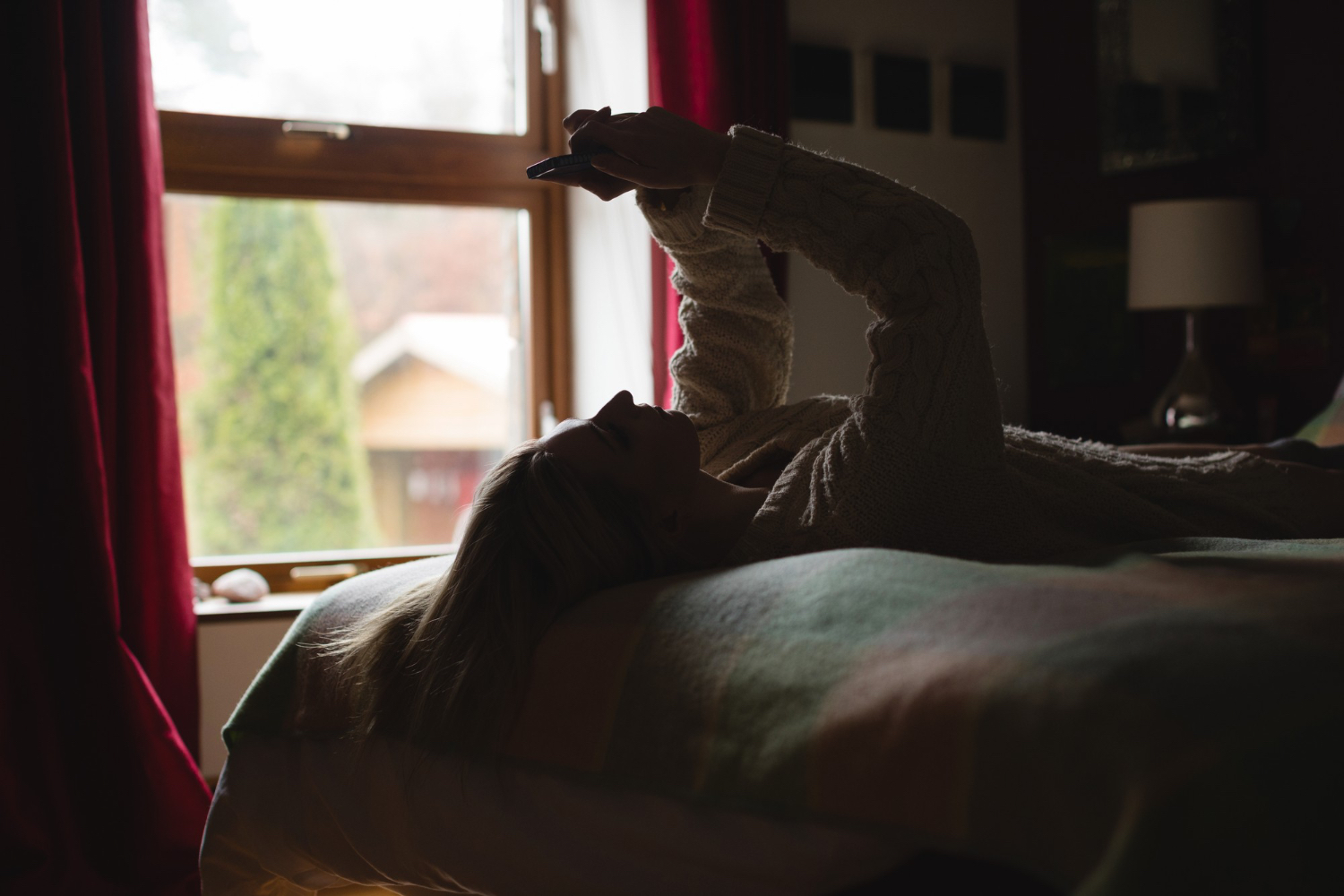 A young woman stays in bed looking at her phone in a dim bedroom, illustrating how lying in bed and staying on screens can make a rest day feel inactive and unstructured.