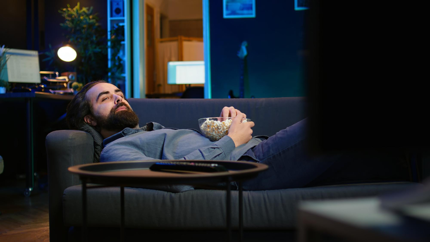 A man lies on a couch at night holding a bowl of popcorn in front of a television, suggesting how snacks, screens, and low activity can push a rest day later into the evening.
