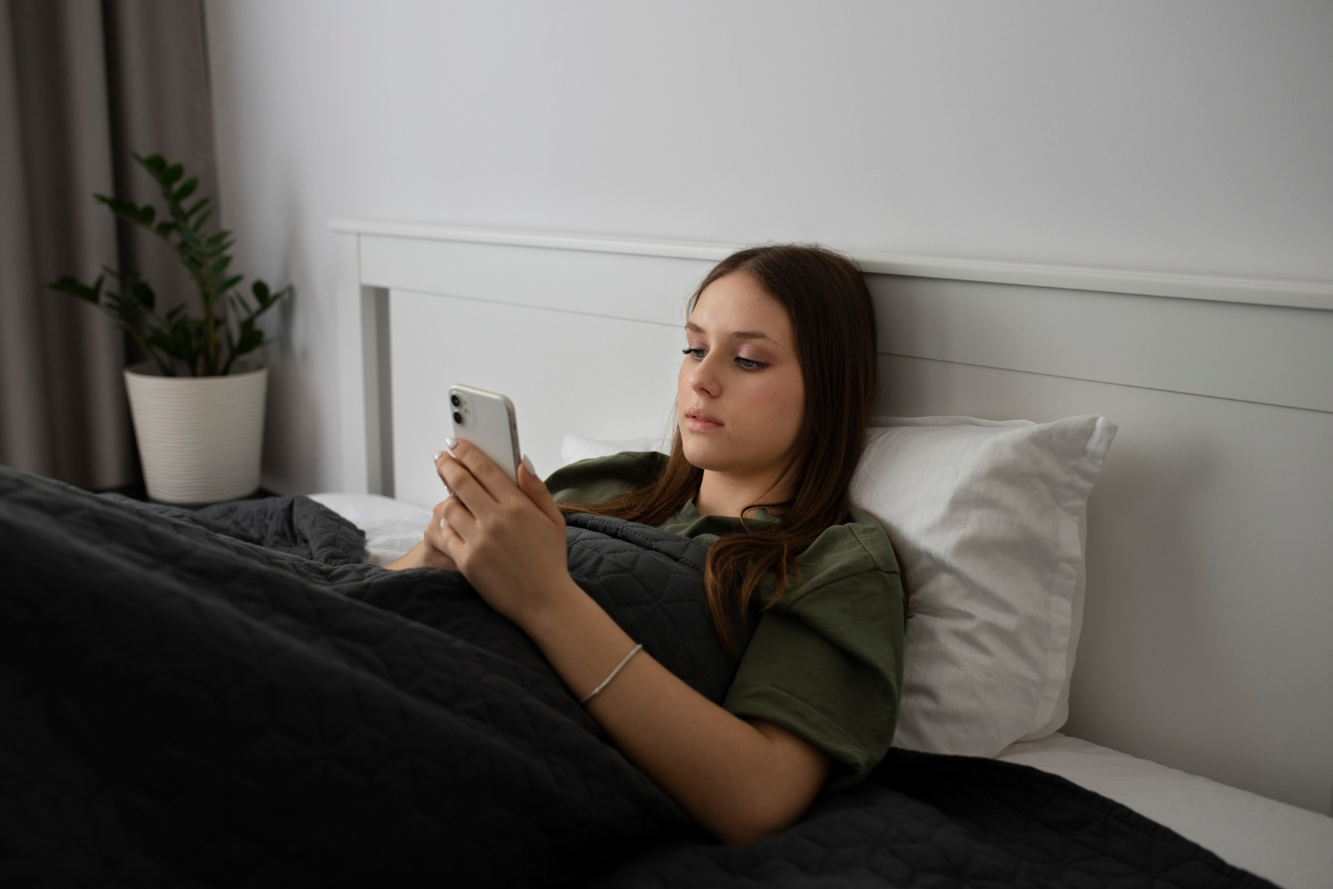 A young woman sits up in bed under a dark blanket while looking at her phone, illustrating screen use and lingering in bed during a slow rest day.