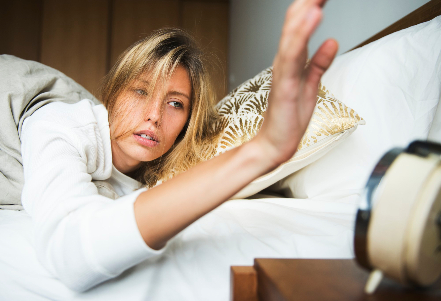 A sleepy woman lies in bed reaching toward an alarm clock with a tired expression.