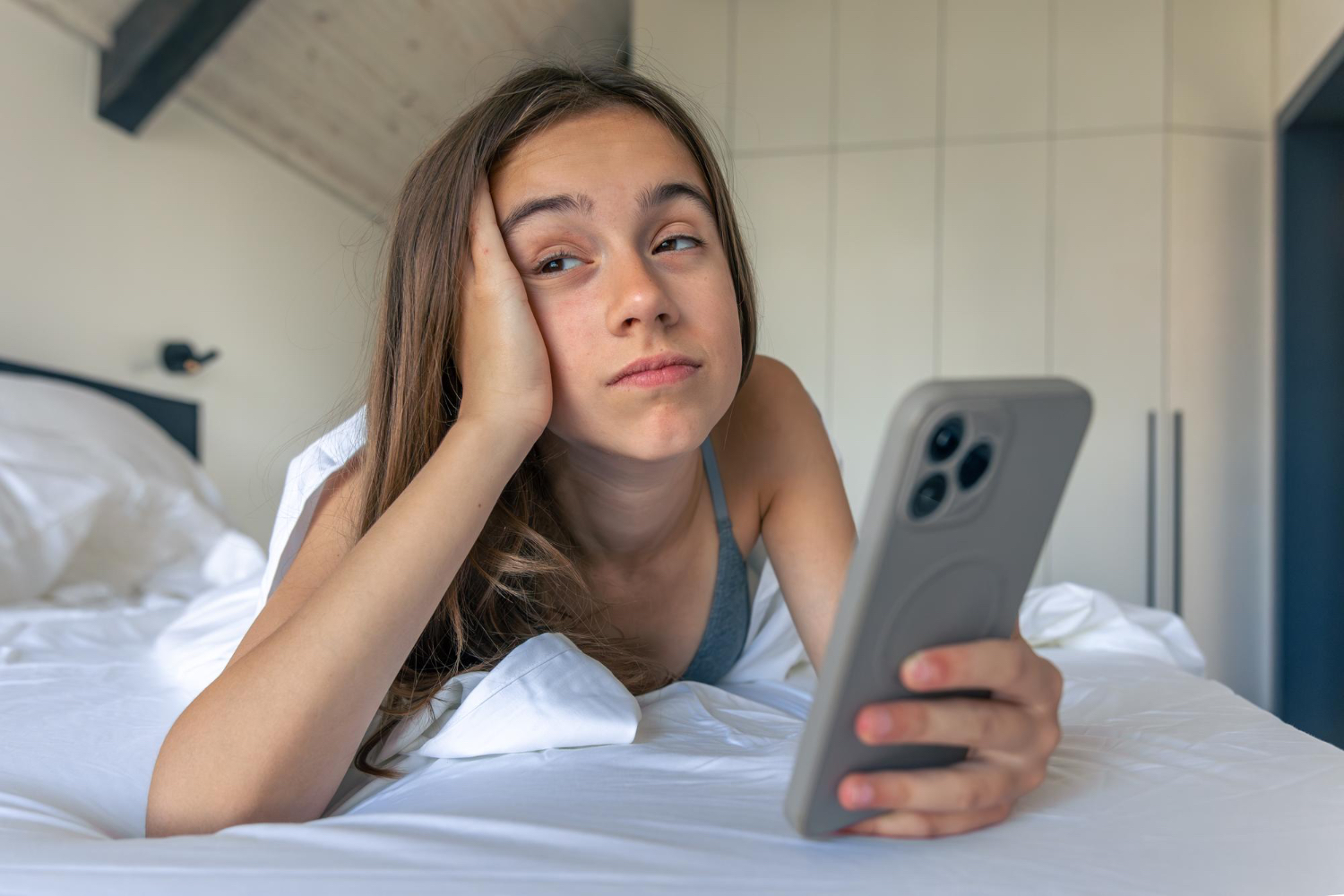 A tired young woman lies on white bedding, staring at her phone with her head resting in her hand.