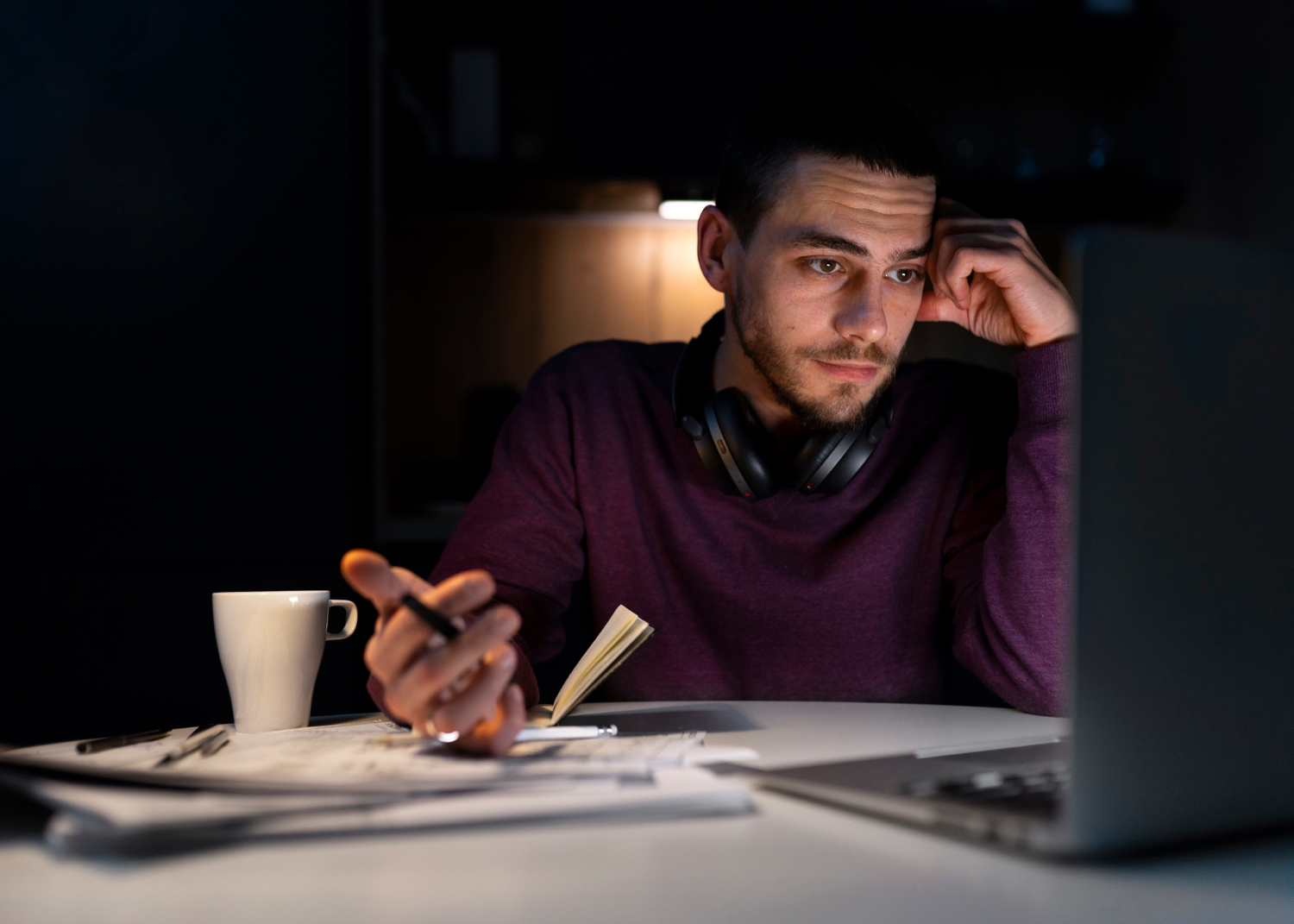 A man works late at a desk in a dark room, looking at a laptop with one hand against his temple. A white mug, scattered papers, a pen, and an open notebook sit on the table, and a pair of headphones rests around his neck.