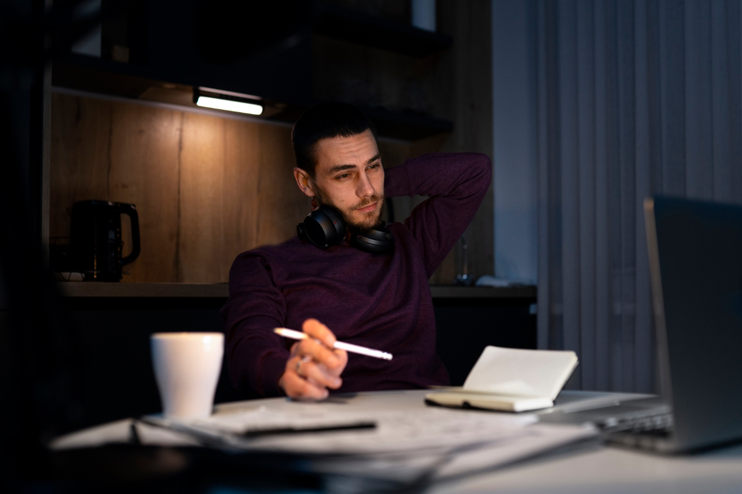 A man sits at a desk late at night with headphones around his neck, leaning back while holding a pen and looking toward a laptop. A white mug, papers, and an open notebook are spread across the desk.