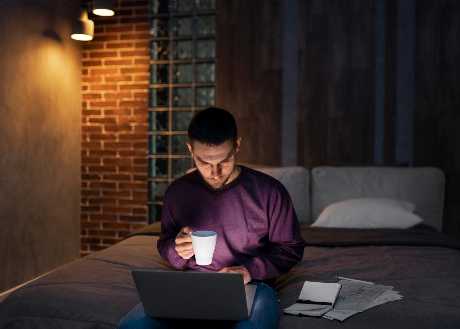 A man sits on a bed at night using a laptop while holding a white mug. Papers and an open notebook lie beside him on the bed, and warm wall lights illuminate the bedroom.