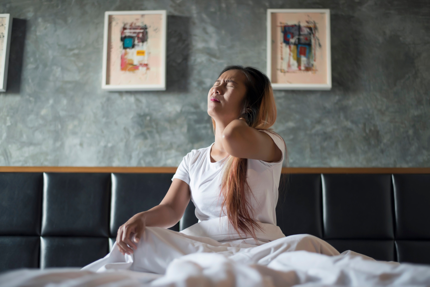 A woman sits up in bed in a white T-shirt, eyes closed and one hand pressed to the back of her neck as if waking with stiffness or fatigue. White bedding, a dark upholstered headboard, and framed artwork are visible behind her.