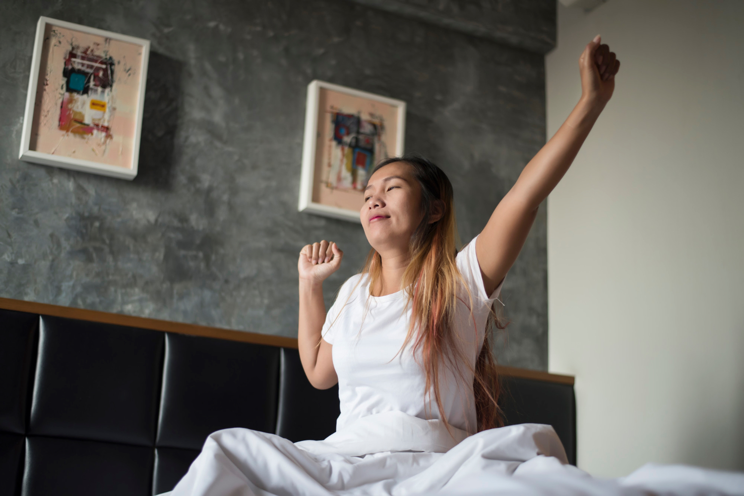 A woman sits up in bed in the morning and stretches both arms, with one arm raised overhead and the other bent near her shoulder. She is surrounded by white bedding with a dark headboard and framed artwork in the background.